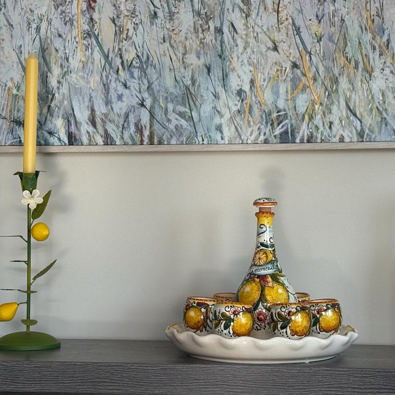 May include: A decorative limoncello set featuring a painted bottle and six matching shot glasses, all adorned with lemon motifs. The set sits on a white, scalloped-edge tray. A yellow candle in a green metal candlestick is on the left.