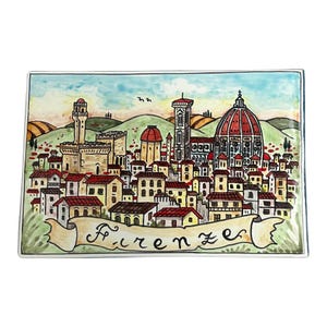 Puede incluir: Un azulejo de cerámica pintado a mano que representa una ciudad colorida de Florencia, Italia. El azulejo presenta una vista panorámica de la ciudad, incluyendo el icónico Duomo, con el texto "Firenze" escrito en una escritura decorativa.