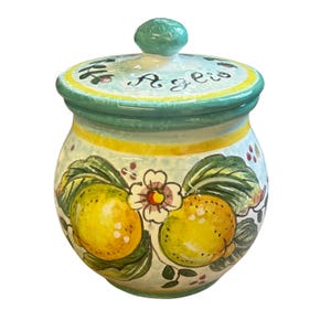 Puede incluir: Un tarro de cerámica con tapa, decorado con un diseño pintado a mano de limones amarillos, hojas verdes y una flor blanca sobre un fondo azul claro. La tapa lleva la palabra Aglio escrita en negro, con un borde en tonos amarillo y azul verdoso.