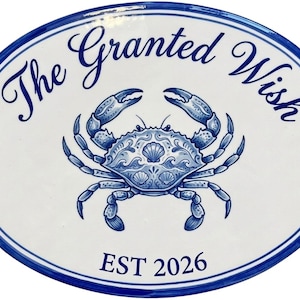 Puede incluir: Una placa ovalada de cerámica blanca con un borde azul y una ilustración central azul de un cangrejo con patrones de conchas detallados. El texto "The Granted Wish" está impreso en azul arriba y "EST 2026" está impreso debajo.