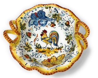 May include: A white ceramic serving bowl with two handles. The bowl is decorated with a rooster, flowers, and leaves in shades of yellow, blue, green, and red. The bowl has a scalloped edge.