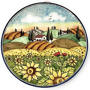 May include: A round ceramic plate with a painted scene of a Tuscan countryside. The plate features a blue sky with white clouds, rolling green hills, a small white house with a red roof, and a cluster of sunflowers in the foreground.