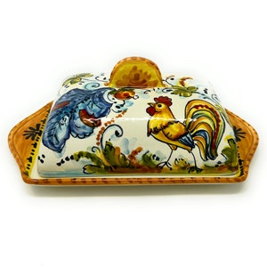 May include: A ceramic butter dish with a rooster and floral design. The dish is white with yellow, blue, green, and red accents. The lid has a handle.