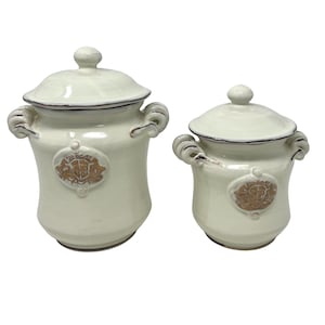 Puede incluir: Dos botes de cerámica blanca con tapas y detalles decorativos dorados. Los botes tienen una forma redondeada y son perfectos para guardar alimentos básicos de la cocina.