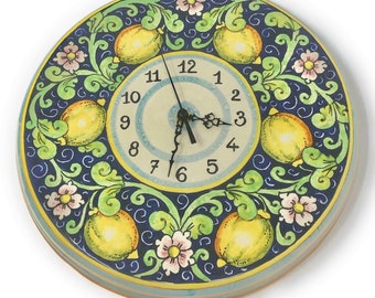 May include: A round wall clock featuring a central cream-colored face with black numerals from 1 to 12 and black ornate hands. The border displays a dark blue background decorated with yellow lemons, light green foliage, and small pink flowers.