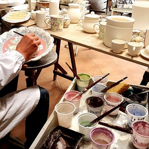 May include: A workshop scene with various ceramic pieces in different stages of completion. A person is painting a decorative plate with a floral design. Paint cups and brushes are arranged on a nearby table, with other finished pottery on shelves.