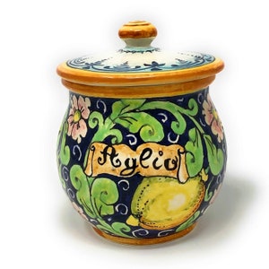 May include: A blue and white ceramic jar with a lid, decorated with green leaves, pink flowers, and a yellow lemon. The jar has the word "Aglio" written on it in a decorative font.