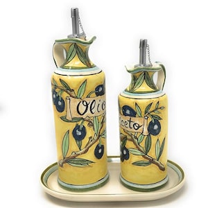 May include: A set of two yellow ceramic olive oil and vinegar bottles with green olive branches and black olives. The bottles have silver spouts and chains. The bottles are sitting on a white oval tray with a green and brown border.