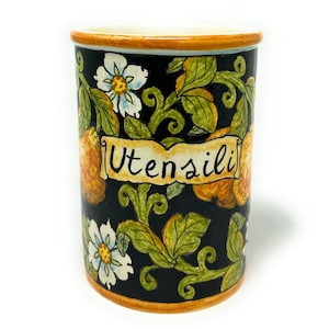 May include: A black ceramic utensil holder with a floral design featuring yellow lemons, white flowers, and green leaves. The holder has a yellow rim and the word "Utensili" written on a yellow banner.