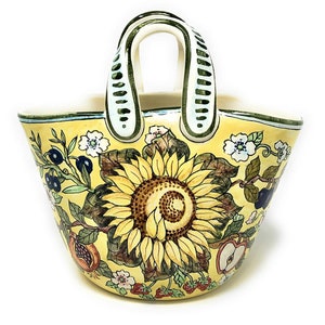 May include: Yellow ceramic basket with a sunflower design. The basket features a large sunflower in the center, surrounded by fruit and floral motifs. The handles are white with green accents. The rim is trimmed in green.