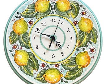 May include: A round ceramic wall clock featuring a hand-painted border of yellow lemons, green leaves, and small white flowers on a light blue background. The center dial has black Arabic numerals from 1 to 12 and two black metal clock hands.