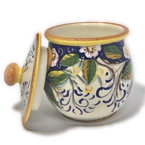 Peut inclure: Un pot en c&eacute;ramique blanche avec un couvercle, d&eacute;cor&eacute; de motifs floraux bleus et jaunes. Le pot a une forme ronde et un petit bouton sur le couvercle.