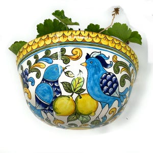 May include: A ceramic wall planter with a vibrant design. The planter features two blue birds, yellow lemons, and green leaves against a white background. The rim is decorated with yellow and green patterns. The planter is filled with green plants.