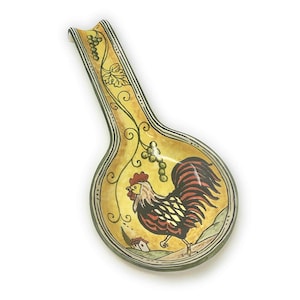 Puede incluir: Un reposacucharas de cerámica con un diseño de gallo. El reposacucharas es amarillo con un borde negro y verde. El gallo es rojo, negro y amarillo. También se representan vides de uva verdes y una pequeña casa.