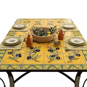 May include: A rectangular dining table with a yellow tiled surface featuring hand-painted olive branch motifs in blue and green. The table is set with four plates, beige placemats, silverware, two terracotta oil cruets, and a woven basket.