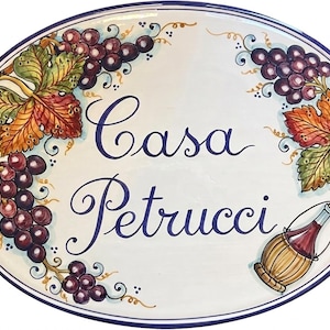 May include: An oval ceramic wall plaque with a white background and a blue border. The text Casa Petrucci is written in blue script in the center. The plaque features hand-painted illustrations of purple grape clusters, green and orange leaves, and a straw-covered wine bottle.