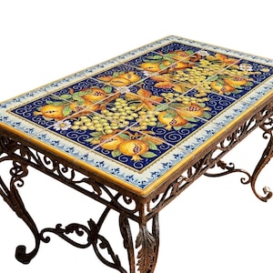 Peut inclure: Une table rectangulaire dotée d'un plateau carrelé orné d'un motif vibrant de grenades jaunes, de feuilles vertes et de raisins blancs sur fond bleu profond, reposant sur une base en fer forgé brun foncé aux pieds courbés.