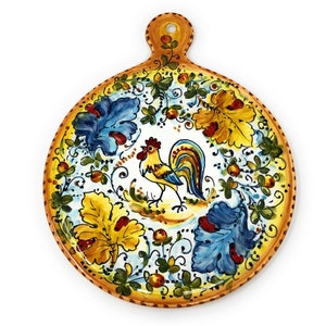 Puede incluir: Un plato de servir redondo de cerámica con un diseño pintado a mano que presenta un gallo, hojas y flores en tonos de azul, amarillo, verde y rojo. El plato tiene un asa en la parte superior.