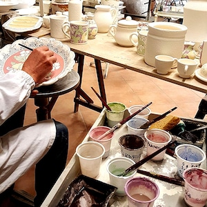 May include: A person is painting a white ceramic plate with a floral design. The plate is on a wooden table with other ceramic pieces, including a jug, a bowl, and a cup. There are also several small containers of paint and brushes on a table.