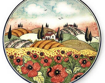 May include: A ceramic plate with a hand-painted design of a Tuscan landscape. The scene features rolling hills, a small white house, and red poppies in the foreground. The plate has a blue rim and a white background.