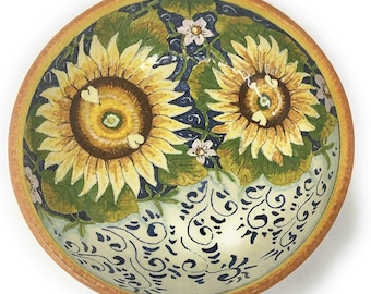 May include: A round ceramic bowl featuring a hand-painted design of two large yellow sunflowers with brown centers, green leaves, and small white flowers against a dark blue background, with a cream-colored base decorated with blue swirls.
