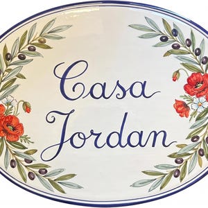 以下が含まれることがあります： 白い背景、青い縁取り、青い筆記体で「Casa Jordan」と書かれた楕円形のセラミックプラーク。 オリーブの枝、オリーブ、赤いポピー、白い花で飾られています。
