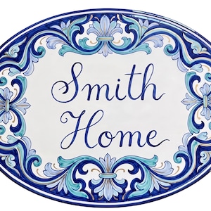 May include: Oval ceramic house plaque with a white center and a decorative blue border. The plaque features the words "Smith Home" in elegant blue script. The border includes floral and scroll designs in shades of blue and teal.