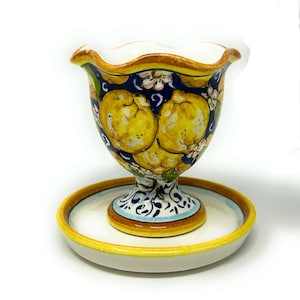 May include: A hand-painted ceramic vase with a matching saucer. The vase features a dark blue background with yellow lemons and white flowers. The rim and saucer have yellow and brown accents. The vase sits on a white surface.