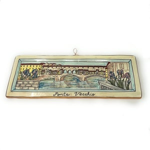 Pode incluir: Um azulejo de cerâmica com uma imagem pintada da Ponte Vecchio em Florença, Itália. A ponte é pintada em tons de marrom, amarelo e branco, com água azul abaixo. O azulejo tem um fundo creme e uma borda azul claro. As palavras "Ponte Vecchio" estão escritas em itálico na parte inferior do azulejo.