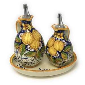 May include: A set of two ceramic oil and vinegar bottles with a lemon design. The bottles have a blue and white background with yellow lemons and green leaves. The bottles are on a white oval tray with a blue and white design. The bottles have silver spouts and chains.