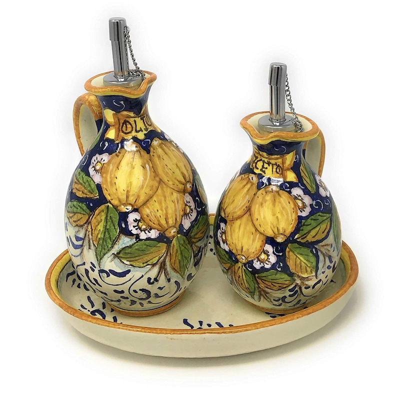 May include: A set of two ceramic oil and vinegar bottles with a lemon design. The bottles have a blue and white background with yellow lemons and green leaves. The bottles are on a white oval tray with a blue and white design. The bottles have silver spouts and chains.