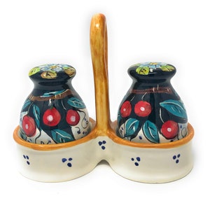 May include: Ceramic salt and pepper shaker set with a handle. The shakers are dark blue with red cherry and green leaf designs. The base is off-white with blue dot accents and an orange trim. The handle is light brown.