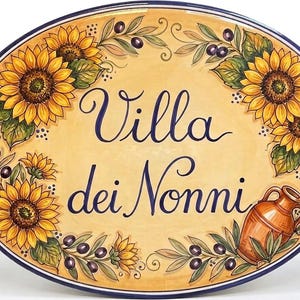 May include: An oval ceramic plaque with a cream-colored background featuring the hand-painted text Villa dei Nonni in dark blue script. The border is decorated with yellow sunflowers, green leaves, dark purple olives, and a terracotta-colored jug.