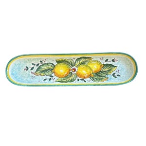 Puede incluir: Una bandeja de servicio ovalada y alargada de cerámica con un diseño pintado a mano de tres limones amarillos rodeados de hojas verdes y una pequeña flor blanca. El fondo es de color azul claro con un borde amarillo y verde azulado.