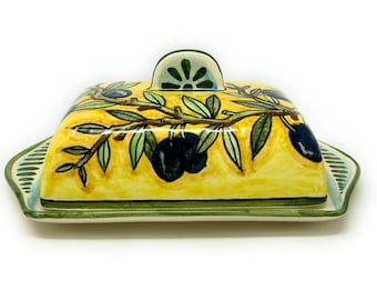 May include: A yellow ceramic butter dish with a green and white olive branch design. The lid has a green knob with a white floral design.