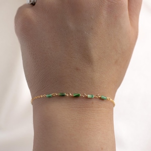 May include: A delicate gold bracelet featuring small, rectangular green beads. The beads are evenly spaced along the gold chain. The bracelet is shown on a person's wrist.