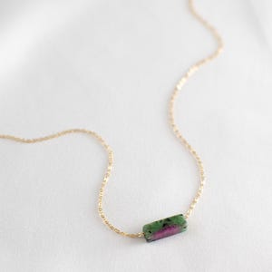 May include: A gold chain necklace with a rectangular gemstone pendant. The pendant is a mix of green, pink, and black. The necklace is displayed on a white background.