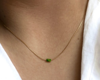 May include: A fine gold chain necklace with a small, rectangular green gemstone pendant. The necklace is shown against a white shirt, contrasting the gold with the vibrant green of the stone. A simple and elegant piece of jewellery.