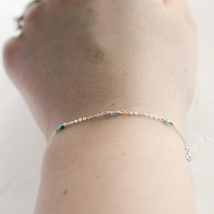 May include: A delicate silver bracelet with small, light blue and teal beads. The bracelet is on a wrist, showcasing the minimalist design and subtle color accents. The chain is fine, adding to the bracelet's elegant simplicity.