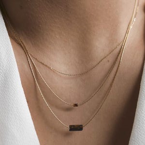 May include: A layered gold necklace set featuring three delicate chains. Each chain has a small, rectangular pendant with a dark brown and gold pattern. The necklaces are displayed against a person's skin and white clothing.