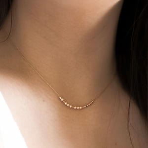May include: A delicate gold necklace with small, round beads and cylindrical accents. The necklace is strung on a fine chain. The necklace is displayed against a person's skin, creating a minimalist and elegant aesthetic.