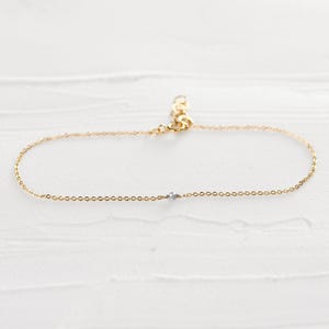 May include: Delicate gold-colored anklet with a small, light blue gemstone. The anklet features a fine chain and an adjustable clasp. The gemstone is centered on the chain. The anklet is displayed on a white surface.