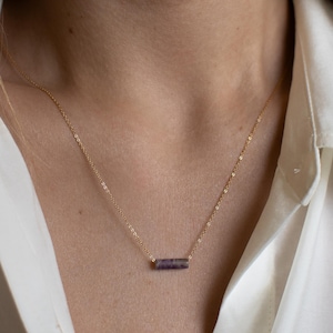 May include: A delicate gold necklace featuring a small, rectangular amethyst pendant. The pendant is a deep purple color with lighter purple stripes. The necklace is worn against a white shirt.