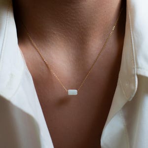 May include: A delicate gold necklace featuring a small, rectangular, pale-colored stone pendant. The necklace is worn against a white collared shirt, creating a minimalist and elegant aesthetic. The chain is fine and the pendant is centered.
