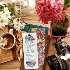 Può includere: Una natura morta con una tazza di caffè e una margherita, un giacinto rosa e una macchina fotografica vintage. Sono presenti anche due segnalibri decorativi e un libro. Il segnalibro recita "Bookmark the Date October Sixth".