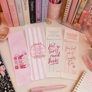 May include: Four rectangular paper bookmarks featuring pink and white designs. Two bookmarks display illustrations of stacked books and bows, while the other two resemble vintage library checkout cards with text reading Hot Girls read Books.