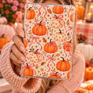 Op de afbeelding: Een doorzichtige tablethoes met een herhalend patroon van oranje en witte pompoenen, roze linten, kleine bloemen en gouden steraccenten op een crèmekleurige achtergrond. Het ontwerp bevat verspreide herfstbladeren en bessen.