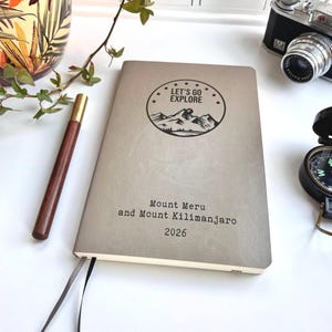 May include: A gray journal with the text "LET'S GO EXPLORE" above a mountain graphic. Below, it reads "Mount Meru and Mount Kilimanjaro 2026." A brown pen and a vintage camera are visible.
