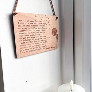 May include: A wooden sign with a light brown background and a white inscription. The inscription is a poem about wishes for difficult days, smiles, rainbows, laughter, sunsets, hugs, beauty, friendships, faith, confidence, courage, patience, and love. The sign is hanging from a brown leather cord.