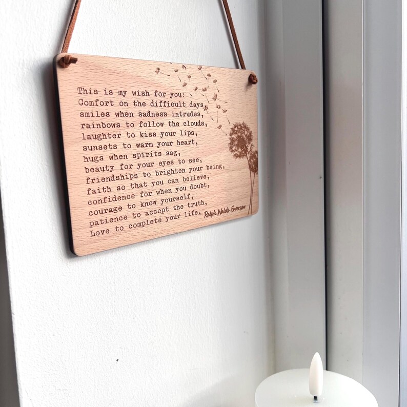 May include: A wooden sign with a light brown background and a white inscription. The inscription is a poem about wishes for difficult days, smiles, rainbows, laughter, sunsets, hugs, beauty, friendships, faith, confidence, courage, patience, and love. The sign is hanging from a brown leather cord.